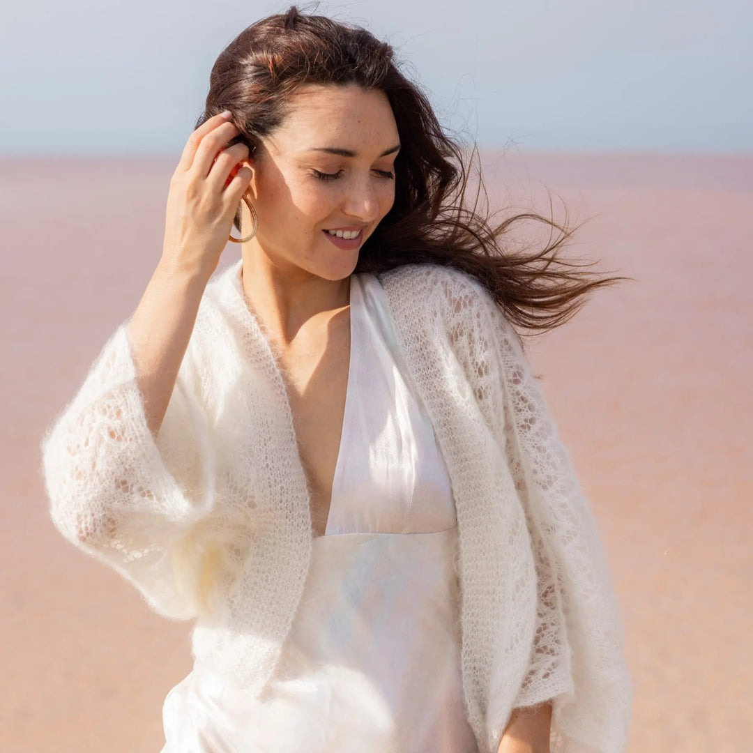GILET MARIÉE EN MOHAIR & SOIE - TRICOTÉ MAIN BOHÈME CHIC - ANNABELLE  couleur ivoire mariage portée par femme brune vue de face sur une plage - Adepte en collaboration avec Mas accessoires