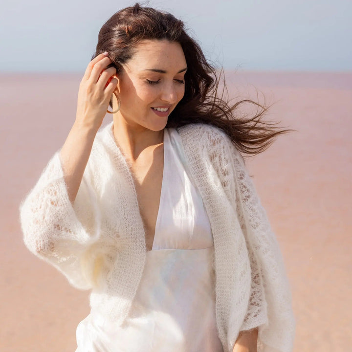 GILET MARIÉE EN MOHAIR & SOIE - TRICOTÉ MAIN BOHÈME CHIC - ANNABELLE  couleur ivoire mariage portée par femme brune vue de face sur une plage - Adepte en collaboration avec Mas accessoires
