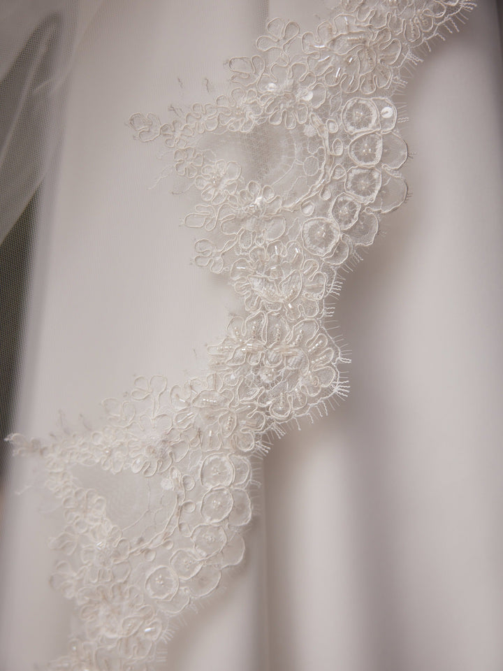 Détail de la bordure en dentelle cordonnée semée de perles nacrées du voile de mariée S564 Bianco Evento